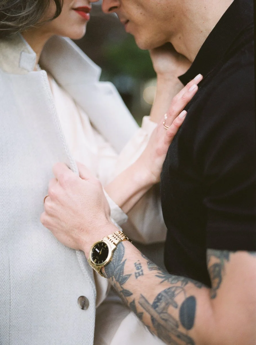 Detail of couple’s hands and clothing, woman touching man’s chest, engagement ring visible, man wearing gold watch