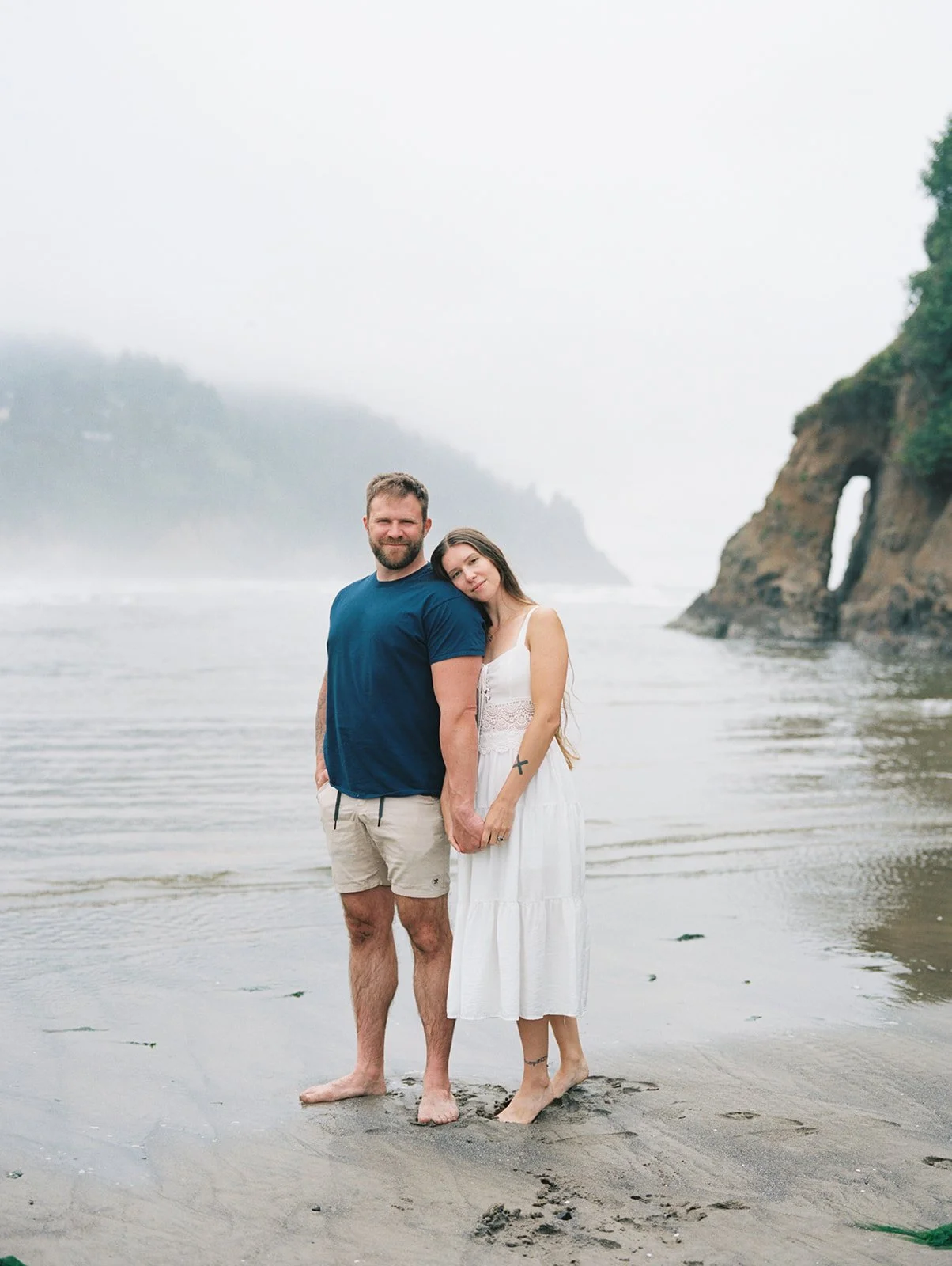 Couple standing barefoot on wet sand near coastal rock arch with misty ocean backdrop.