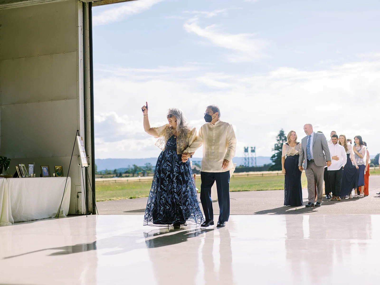 Older couple making their grand entrance into the Pearson Air Museum hangar, the woman in a navy floral gown waving triumphantly
