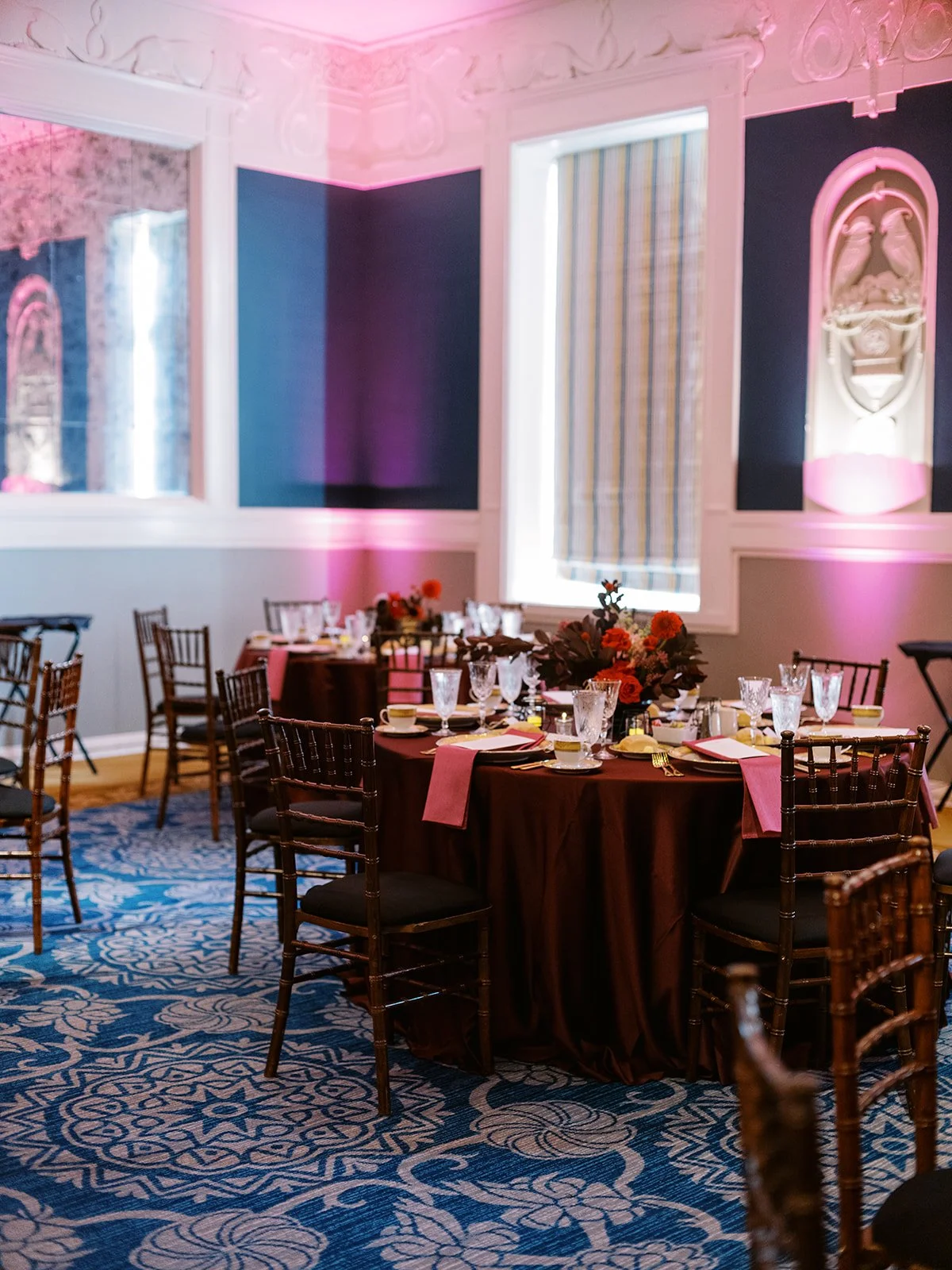 Reception tables set with burgundy linens, glassware, and floral centerpieces under pink uplighting.