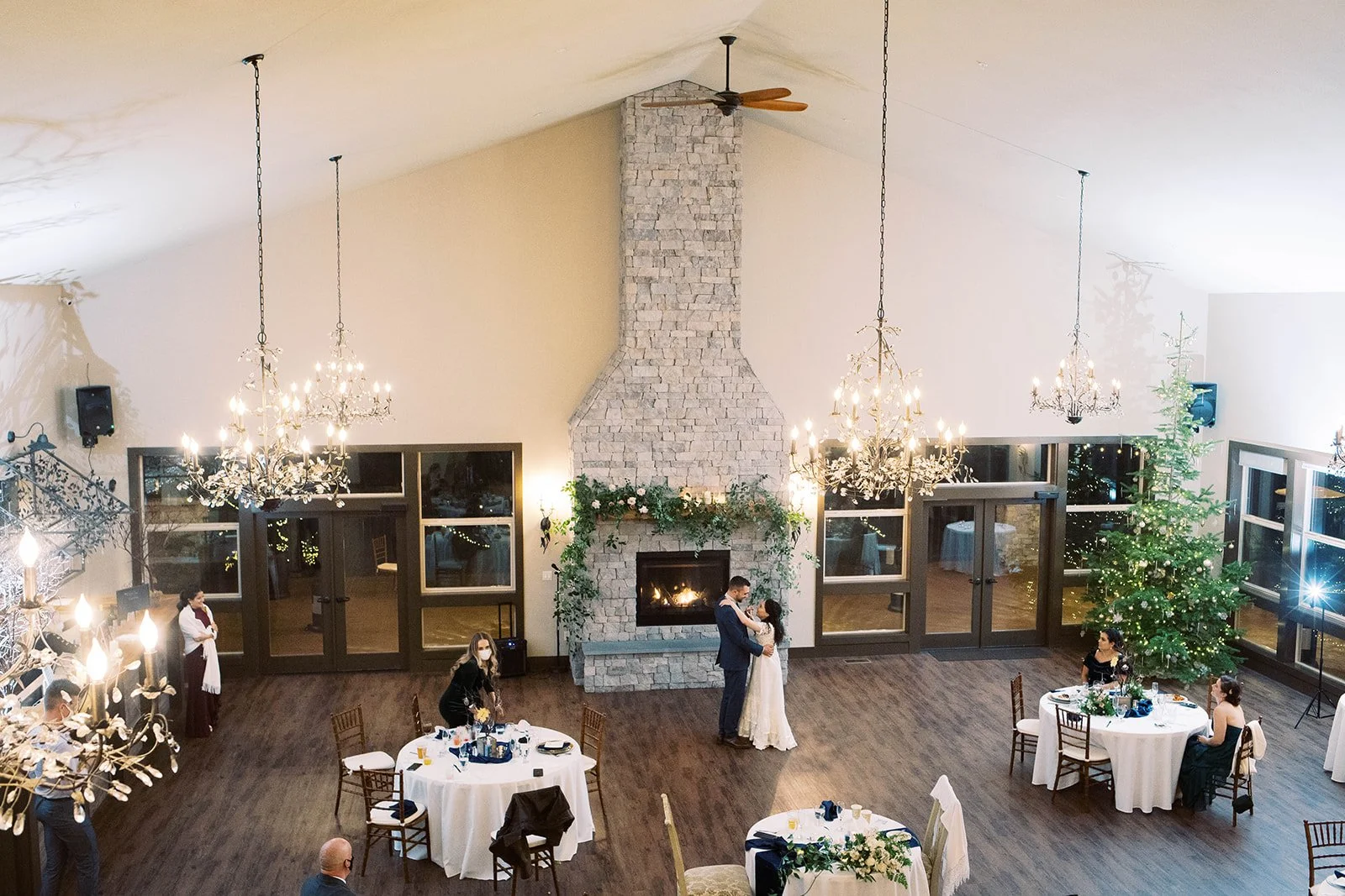 Bride and groom dance together in an intimate reception space with chandeliers, fireplace, and Christmas tree.
