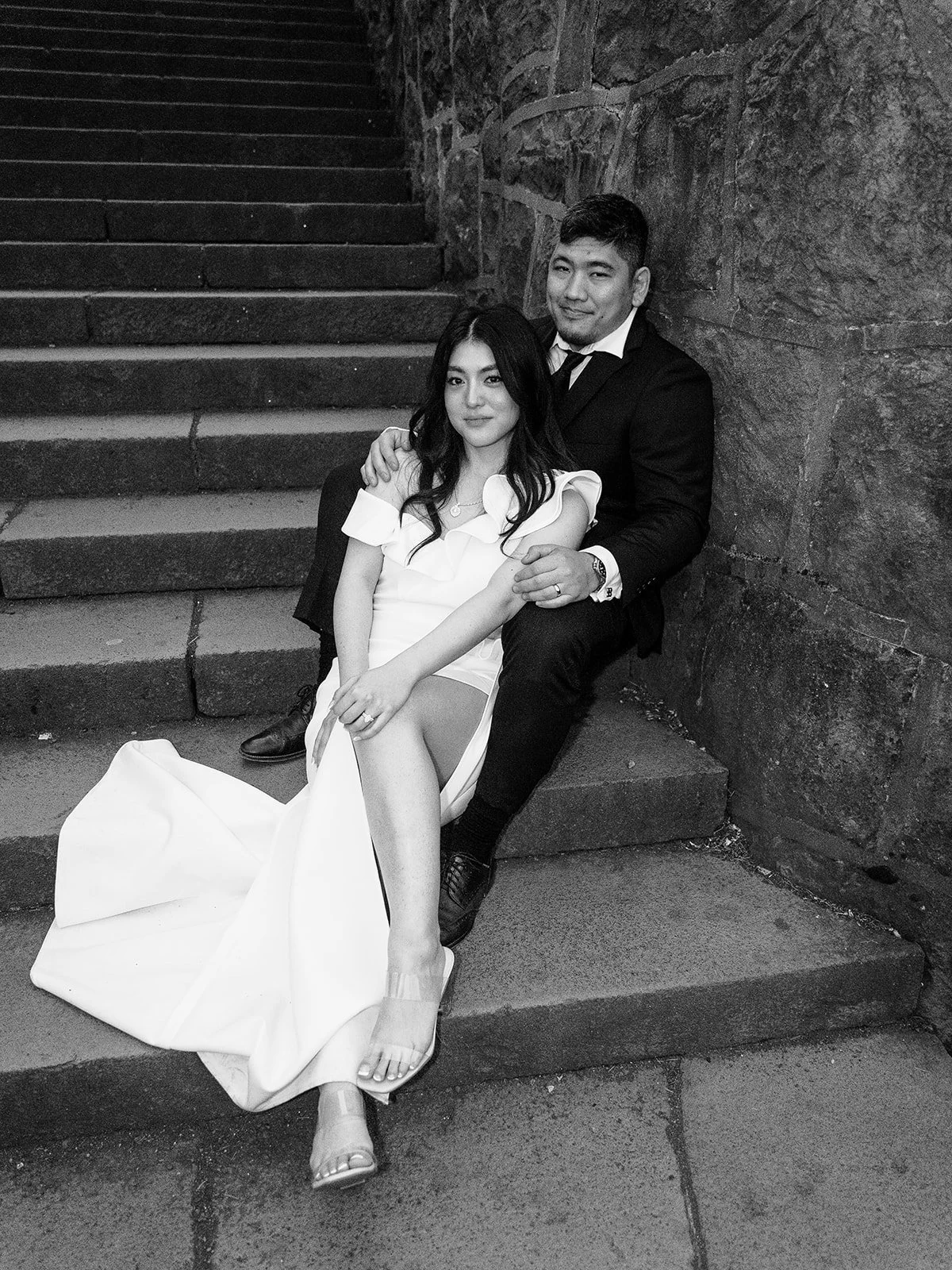 Black and white photo of the bride and groom seated together on the stone steps at The Grotto's upper overlook, her white slit gown pooling around her as he wraps his arm around her shoulder