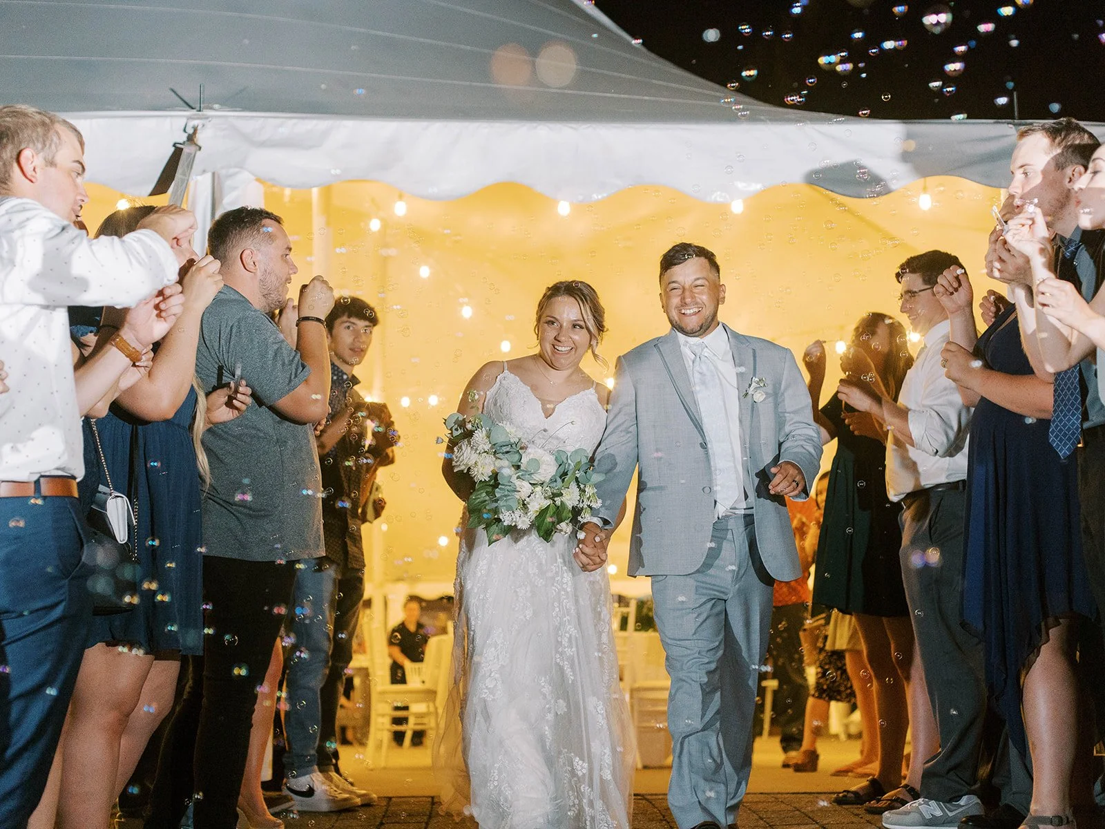 Newlywed couple walks hand in hand through a bubble send-off at night outside the reception tent, surrounded by cheering guests.