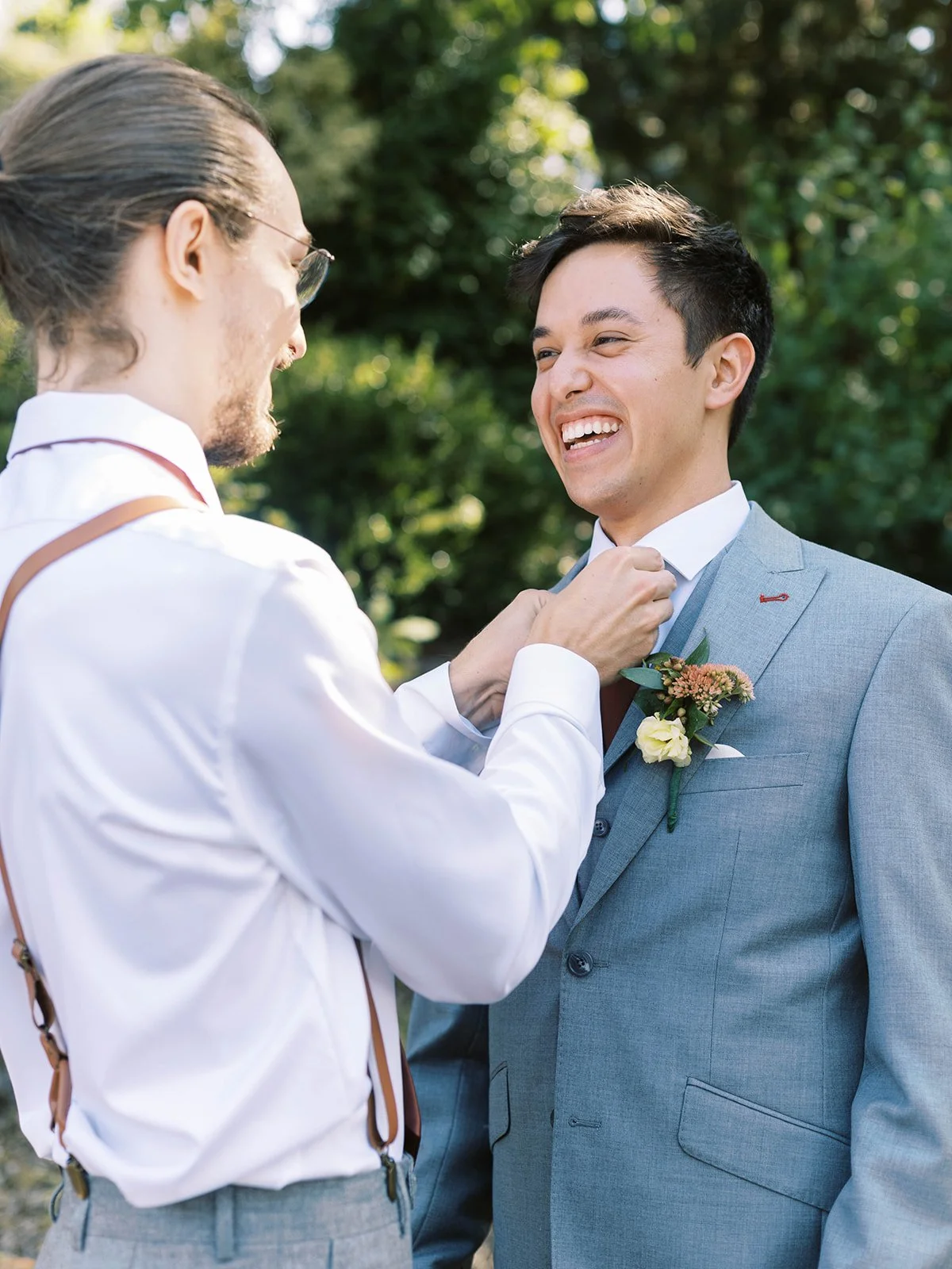 Groom smiling as another man adjusts his tie outdoors, boutonniere visible against a gray suit.