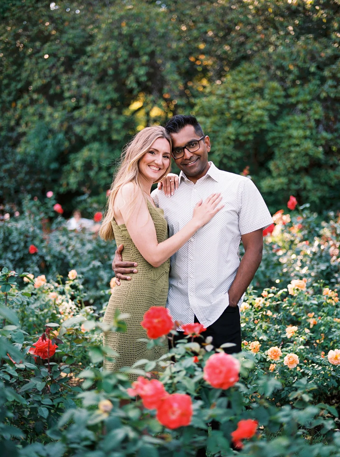 A smiling couple posed together among red and orange roses in a sunlit garden.