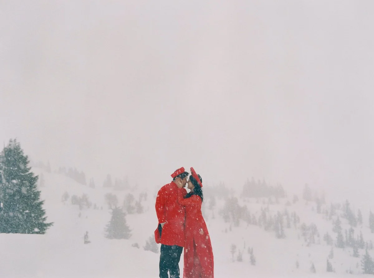 Couple in red Vietnamese áo dài embracing in heavy snowfall at Mount Hood with misty mountain backdrop.