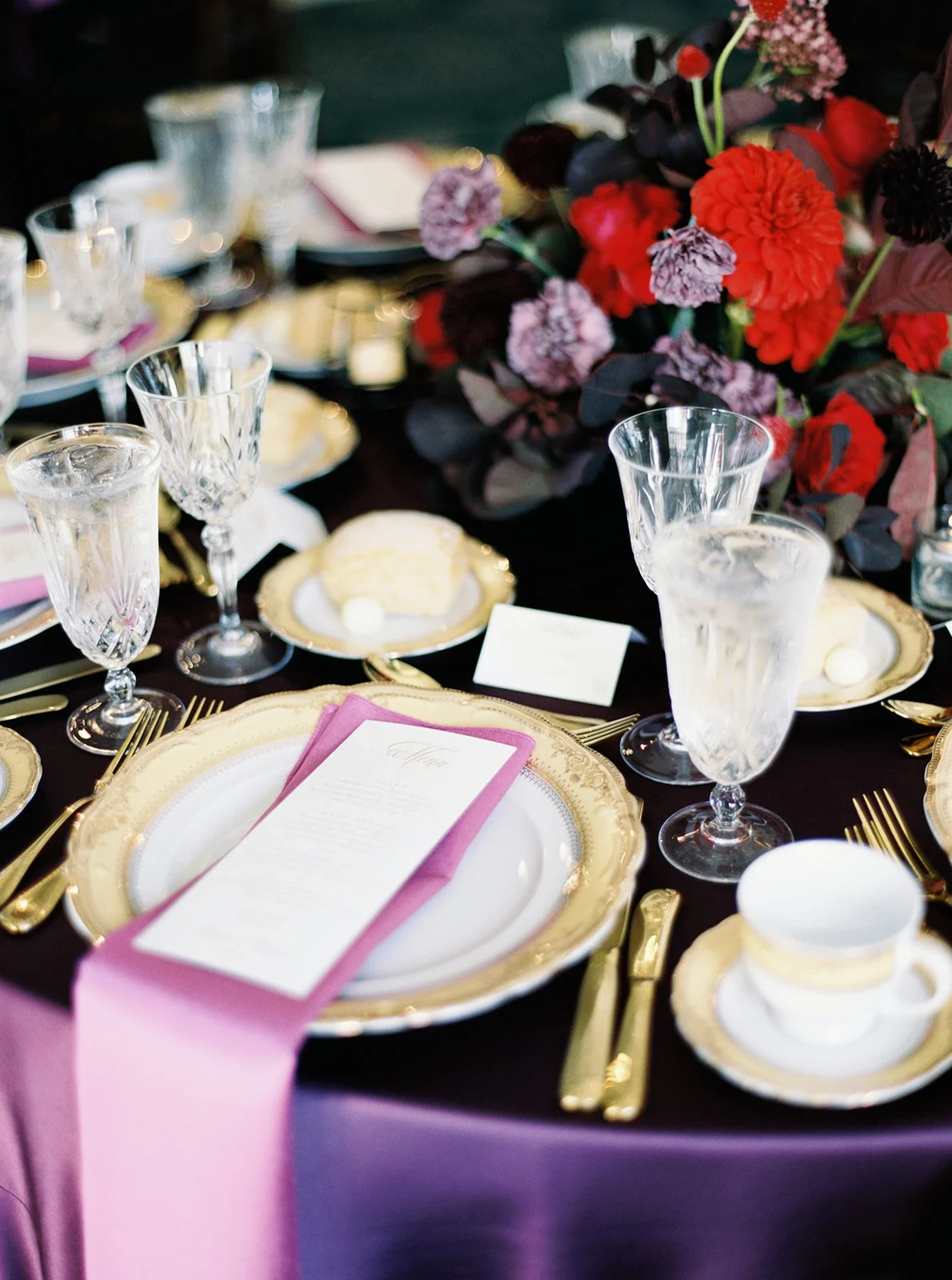 Formal place setting with gold-rimmed china, crystal glassware, and a bold red and burgundy floral centerpiece.