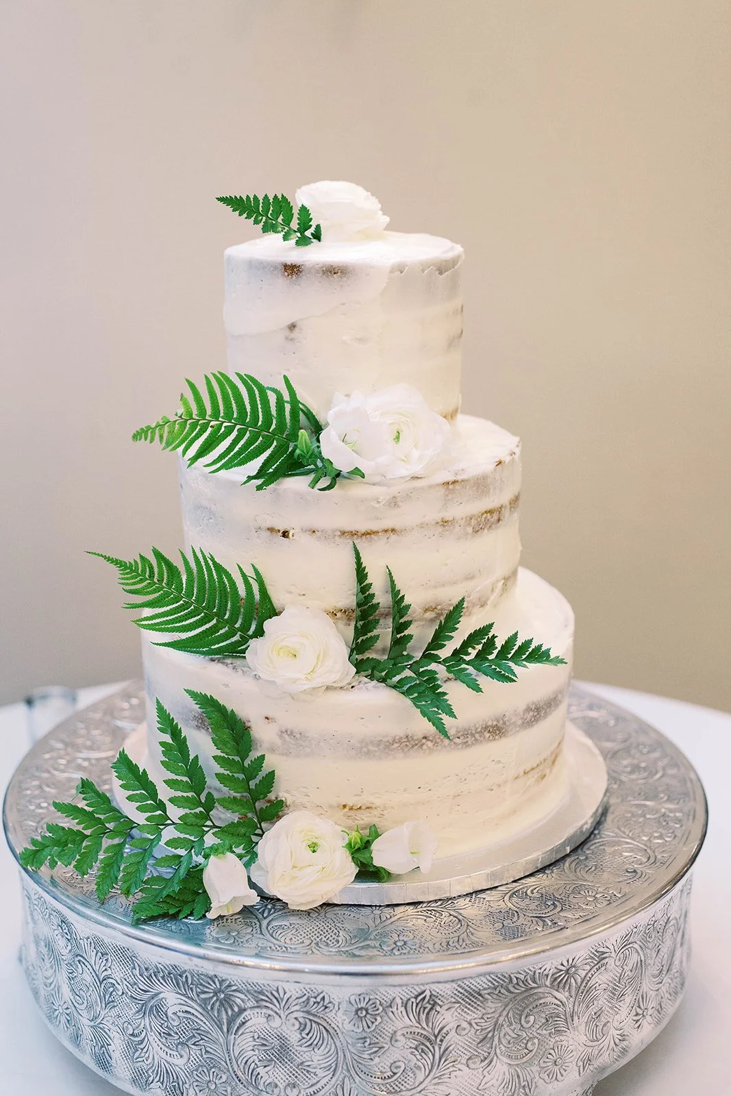 Three-tier semi-naked wedding cake decorated with white flowers and fresh green ferns.