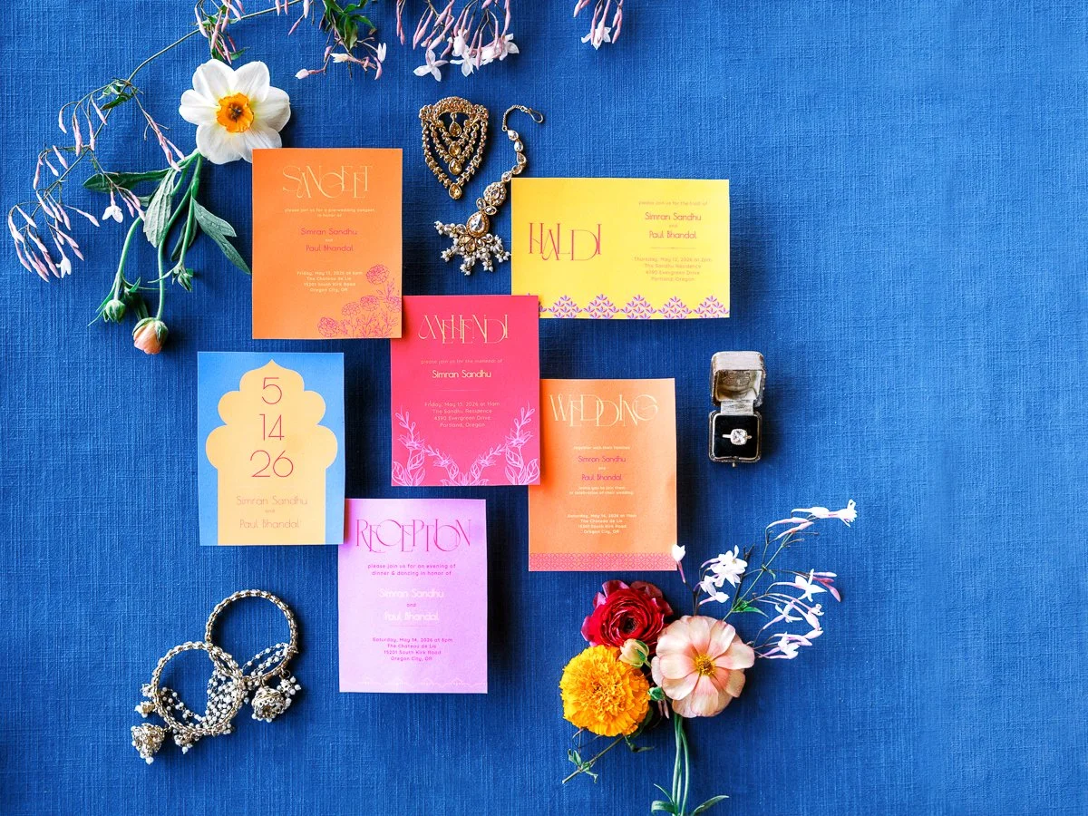 Colorful South Asian wedding invitation suite with floral details, gold jewelry, and engagement ring styled on a royal blue backdrop.