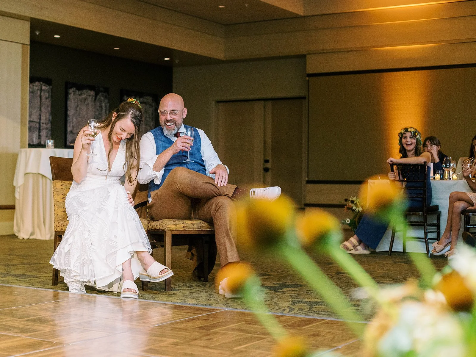 Bride and groom seated on the dance floor laughing with wine glasses during a reception game, with guests watching in the background