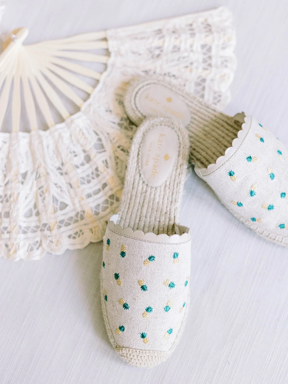 Kate Spade pineapple-embroidered espadrille mules beside a white lace hand fan on a white surface
