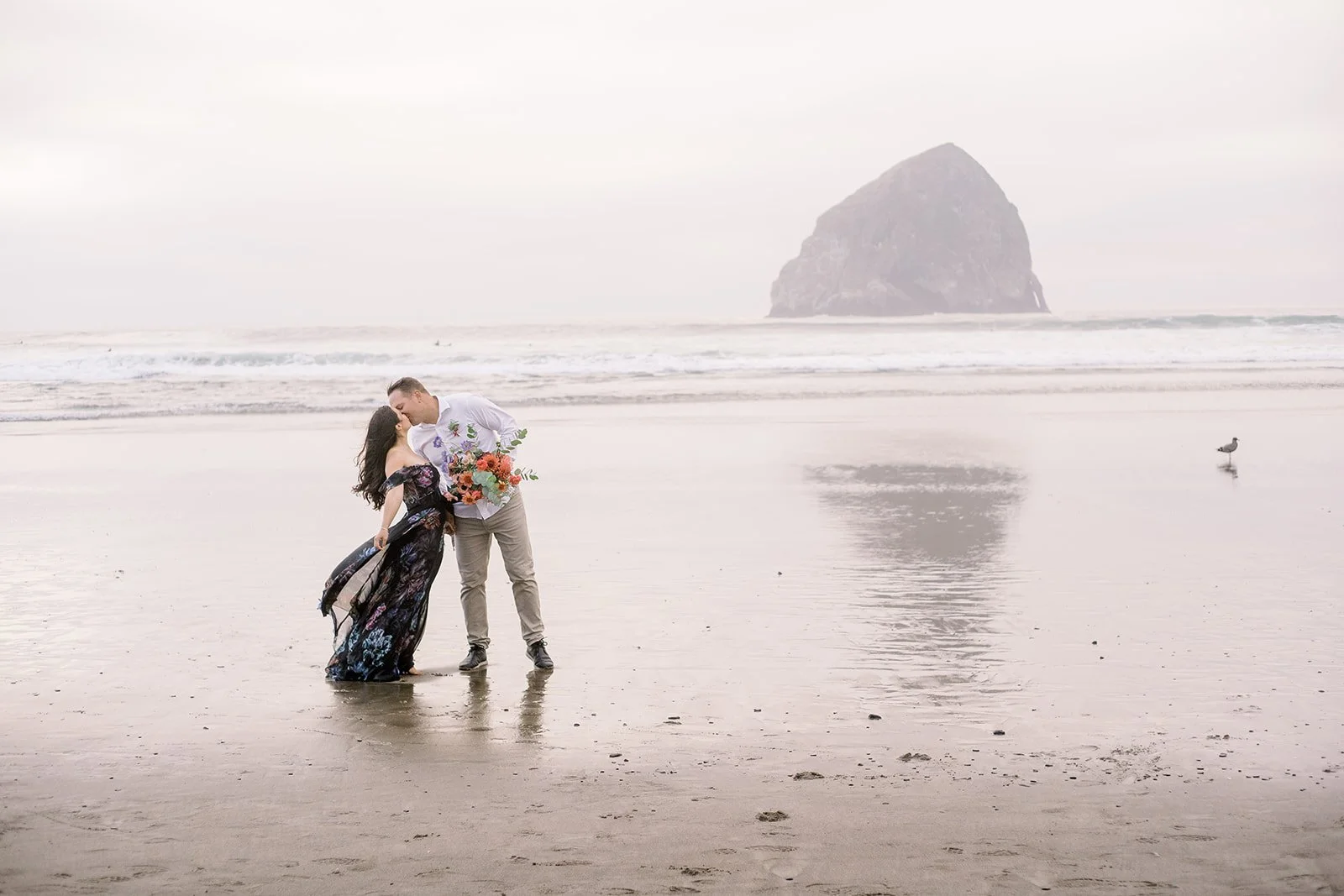 Couple kissing on reflective beach with Haystack Rock in distance, soft foggy Oregon Coast scene