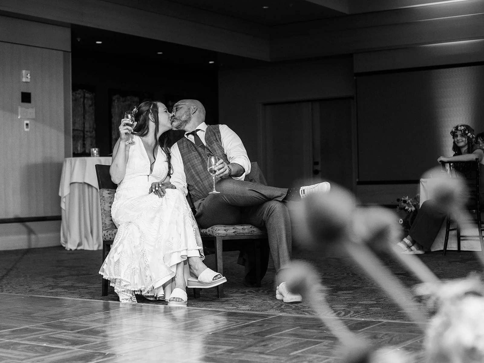 Black and white photo of the bride and groom stealing a kiss while seated during the reception, a flower-crowned guest visible in the background