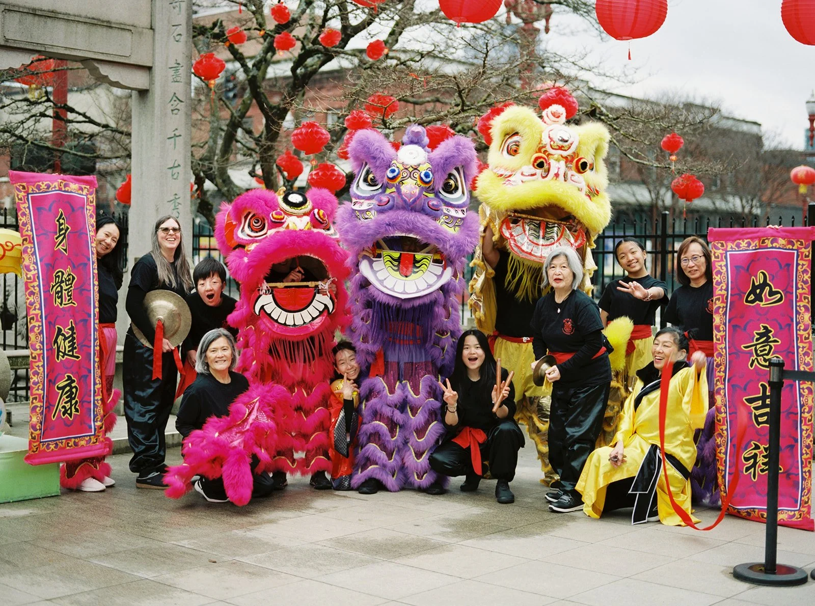 Chinese lion dance performers and musicians pose beneath red lanterns with colorful lion costumes.