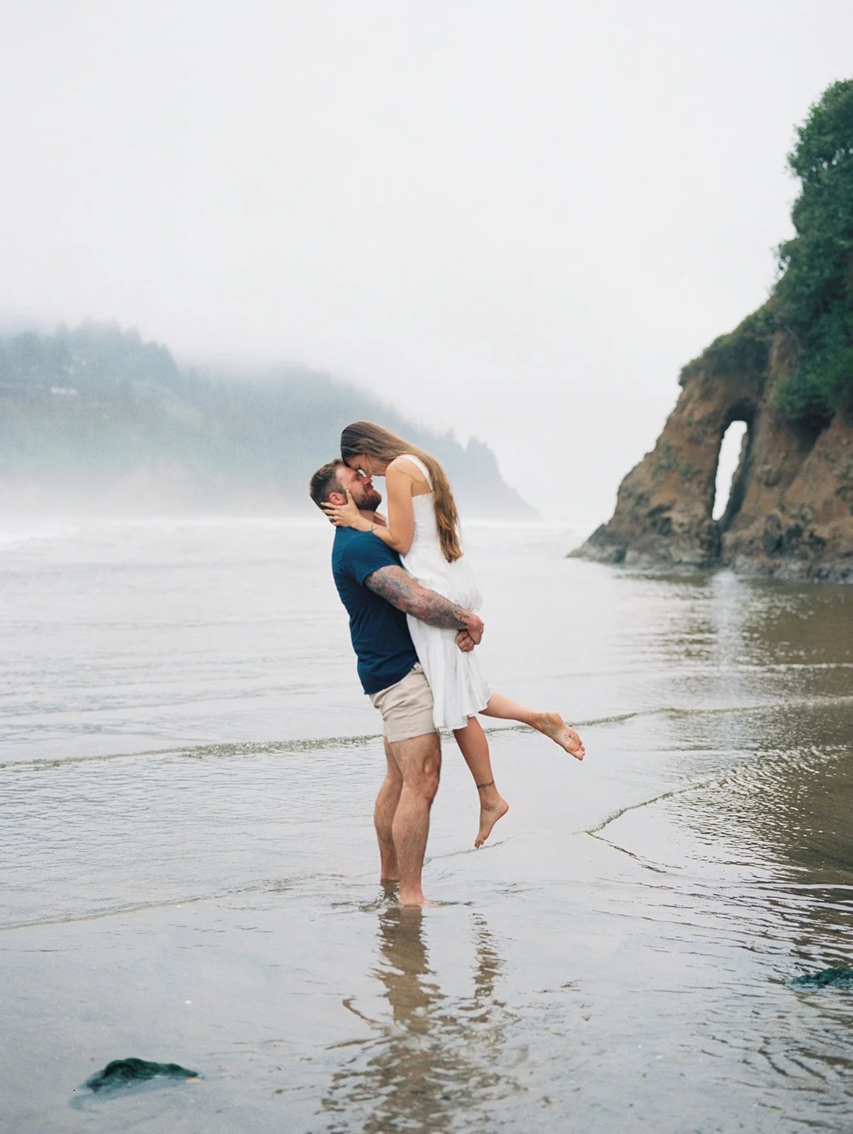 Man lifting woman in white dress in shallow water near rocky arch on foggy beach.