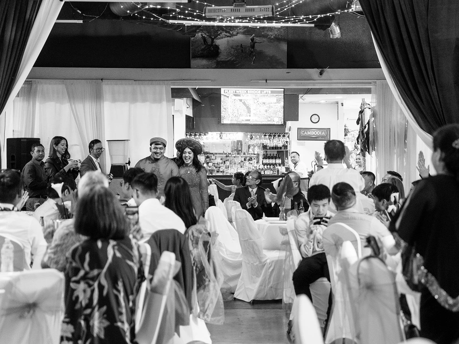 Black and white wide shot of the couple making their grand entrance in red Vietnamese áo dài at Mekong Bistro, guests seated at white-draped tables applauding on either side