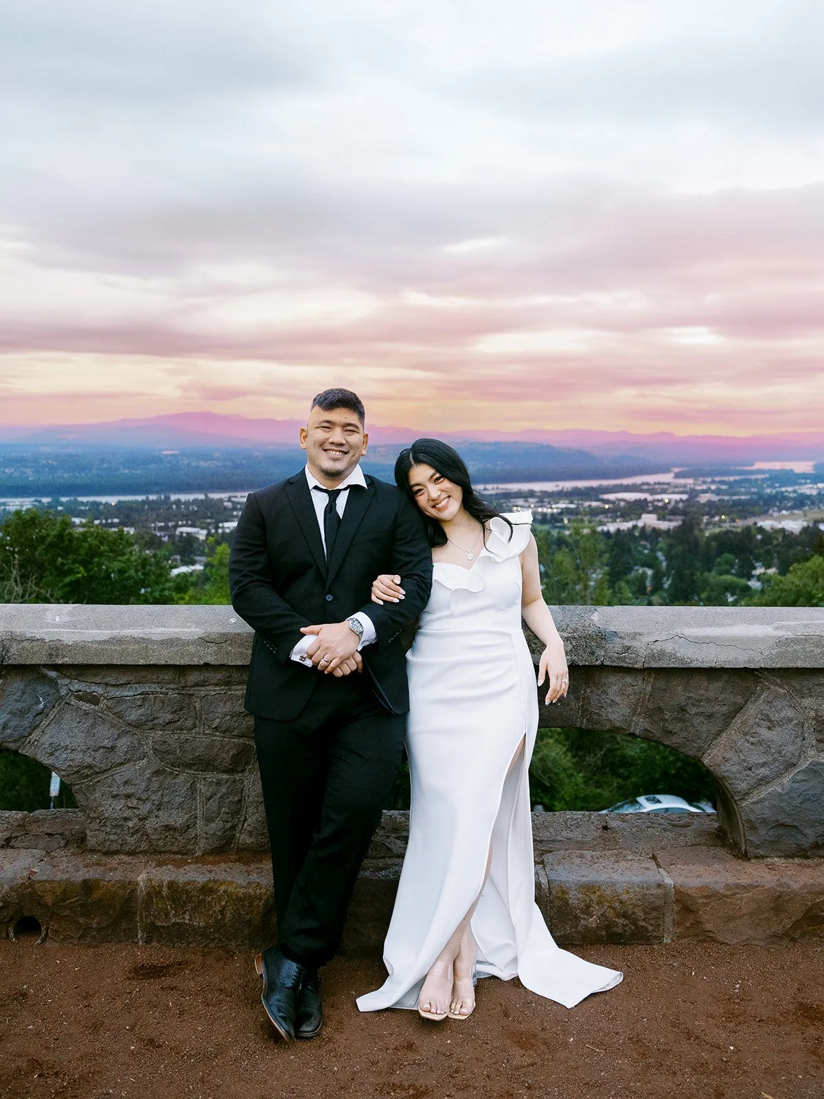 Bride and groom in wedding attire posing against a stone balustrade at The Grotto's upper overlook at sunset, the Columbia River valley and pink-orange sky stretching behind them