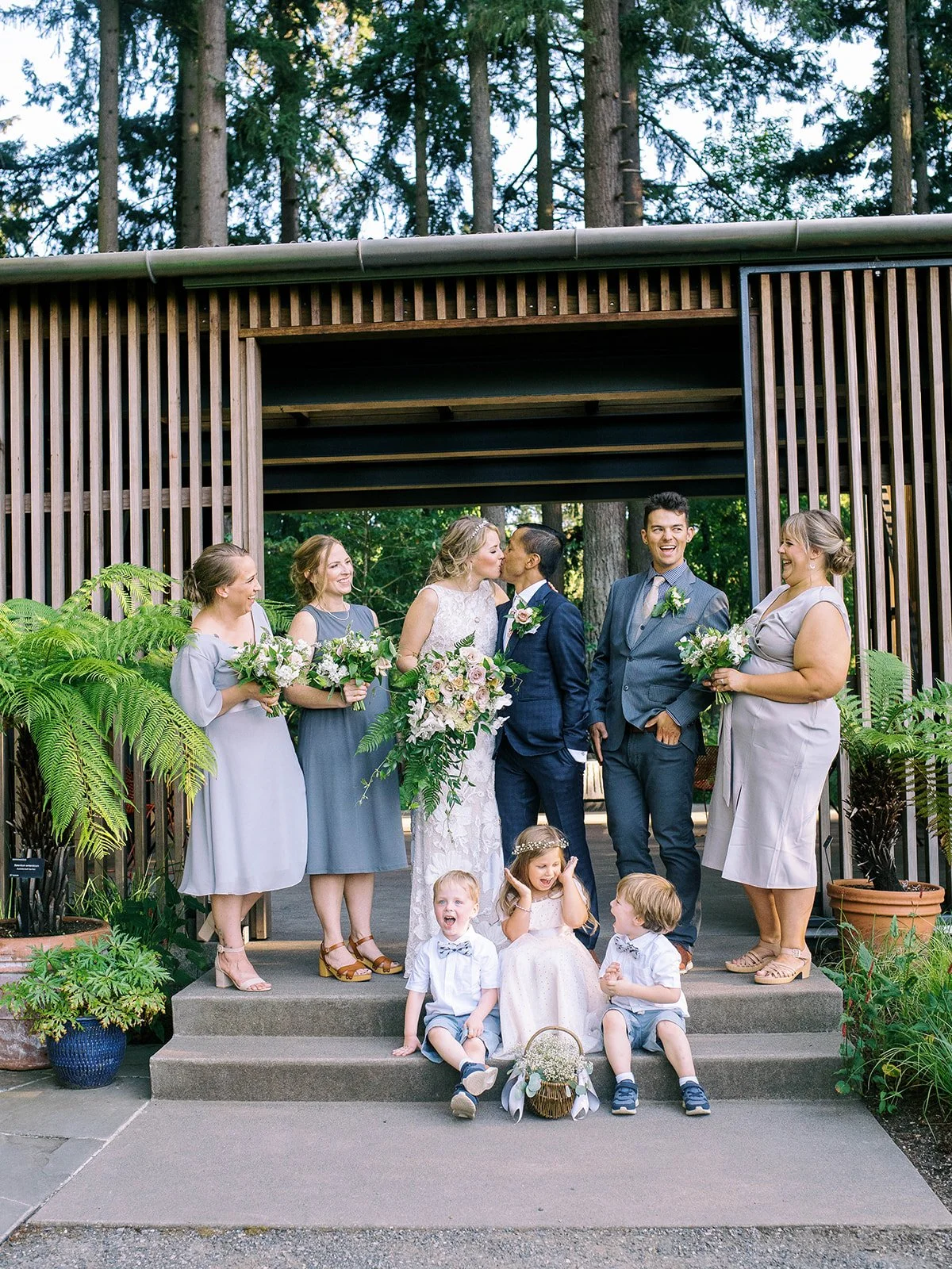 Wedding party portrait in front of modern wood architecture, with three children seated on the steps.