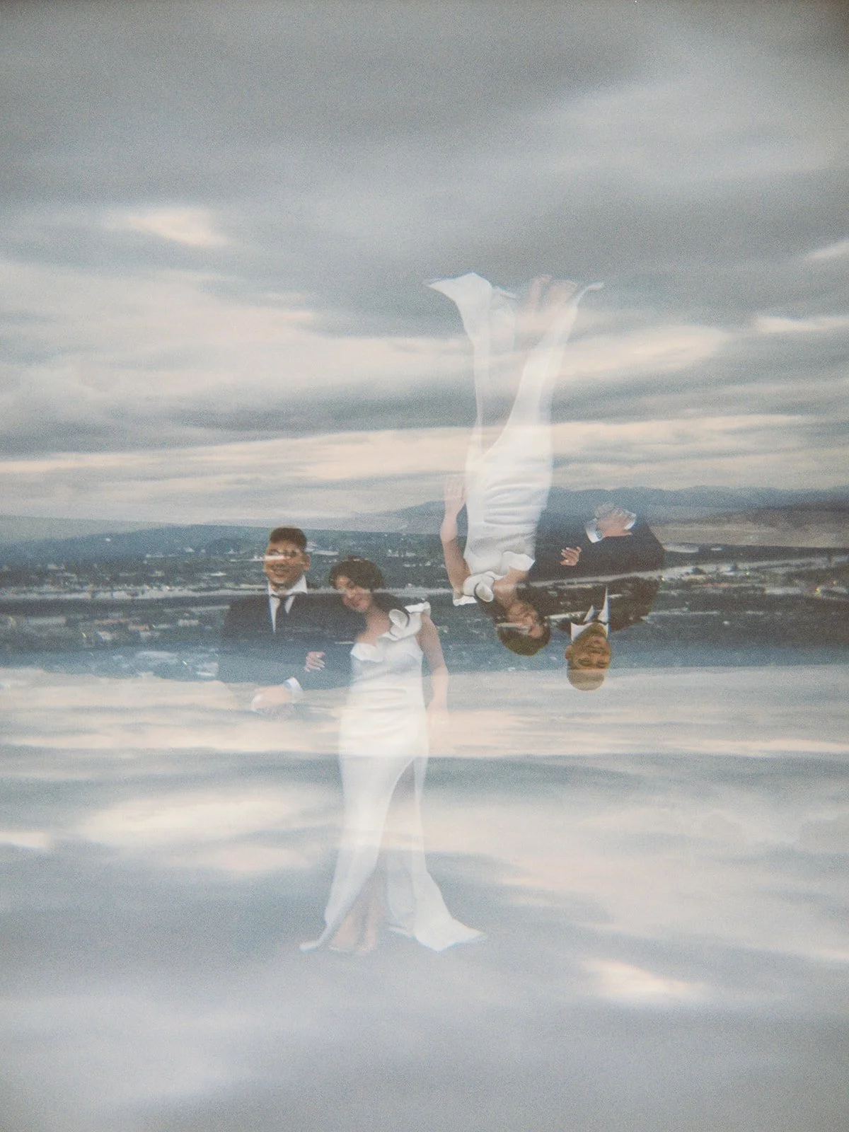 Double-exposure film image of the bride and groom at The Grotto overlook, their figures layered twice against a moody overcast sky and the Portland cityscape below