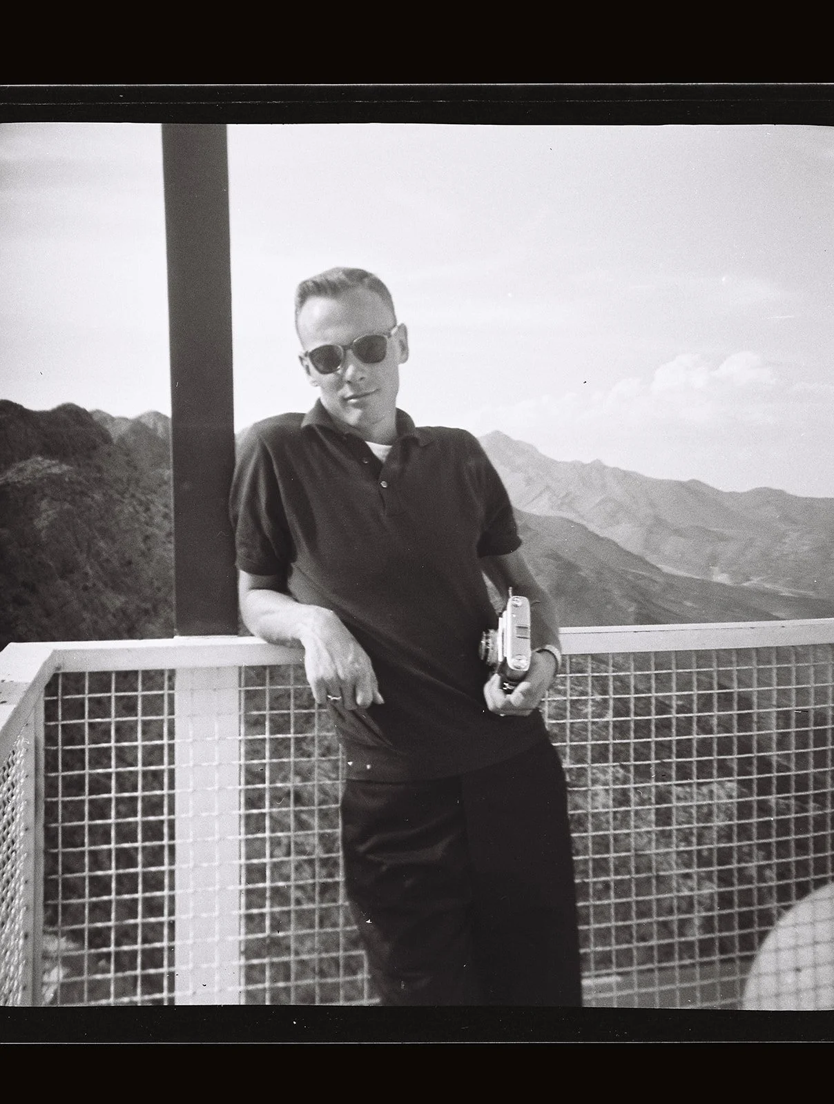 Black-and-white photograph of a man with a camera standing at a mountain overlook.