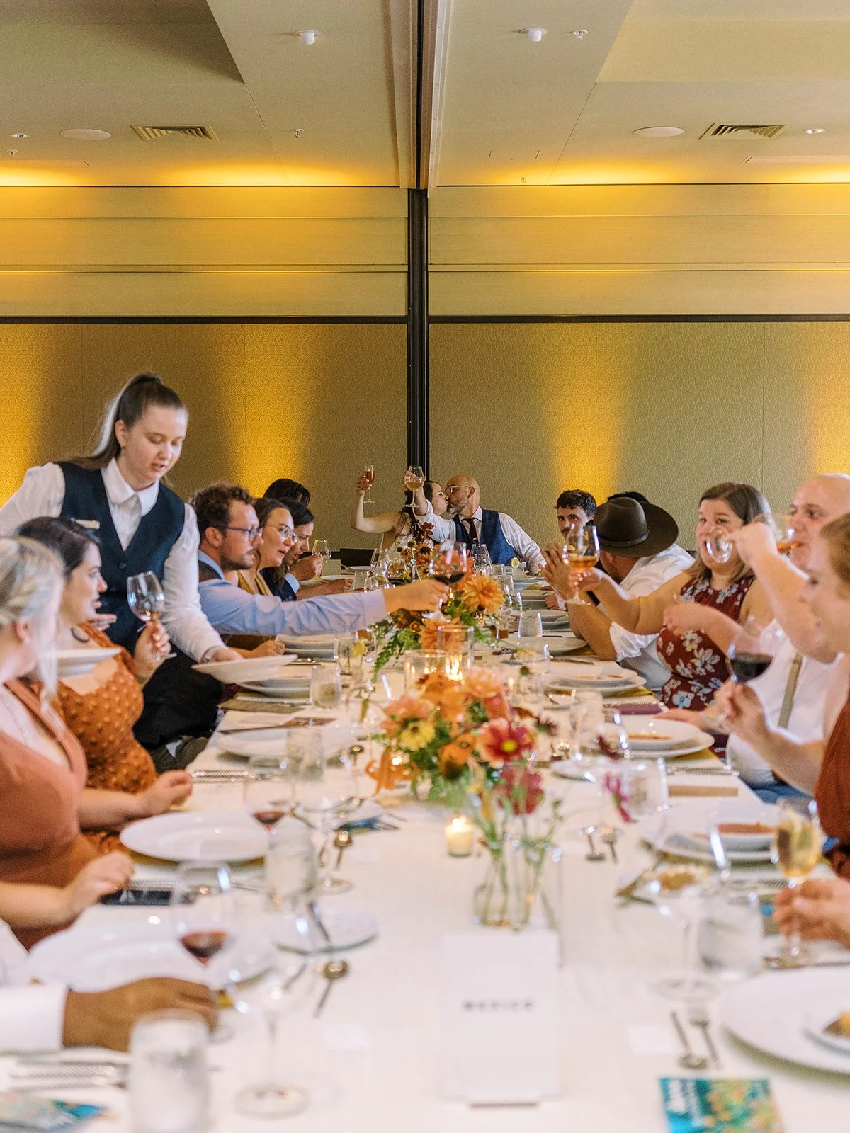 Wedding guests raising glasses around a long reception table decorated with colorful floral centerpieces.