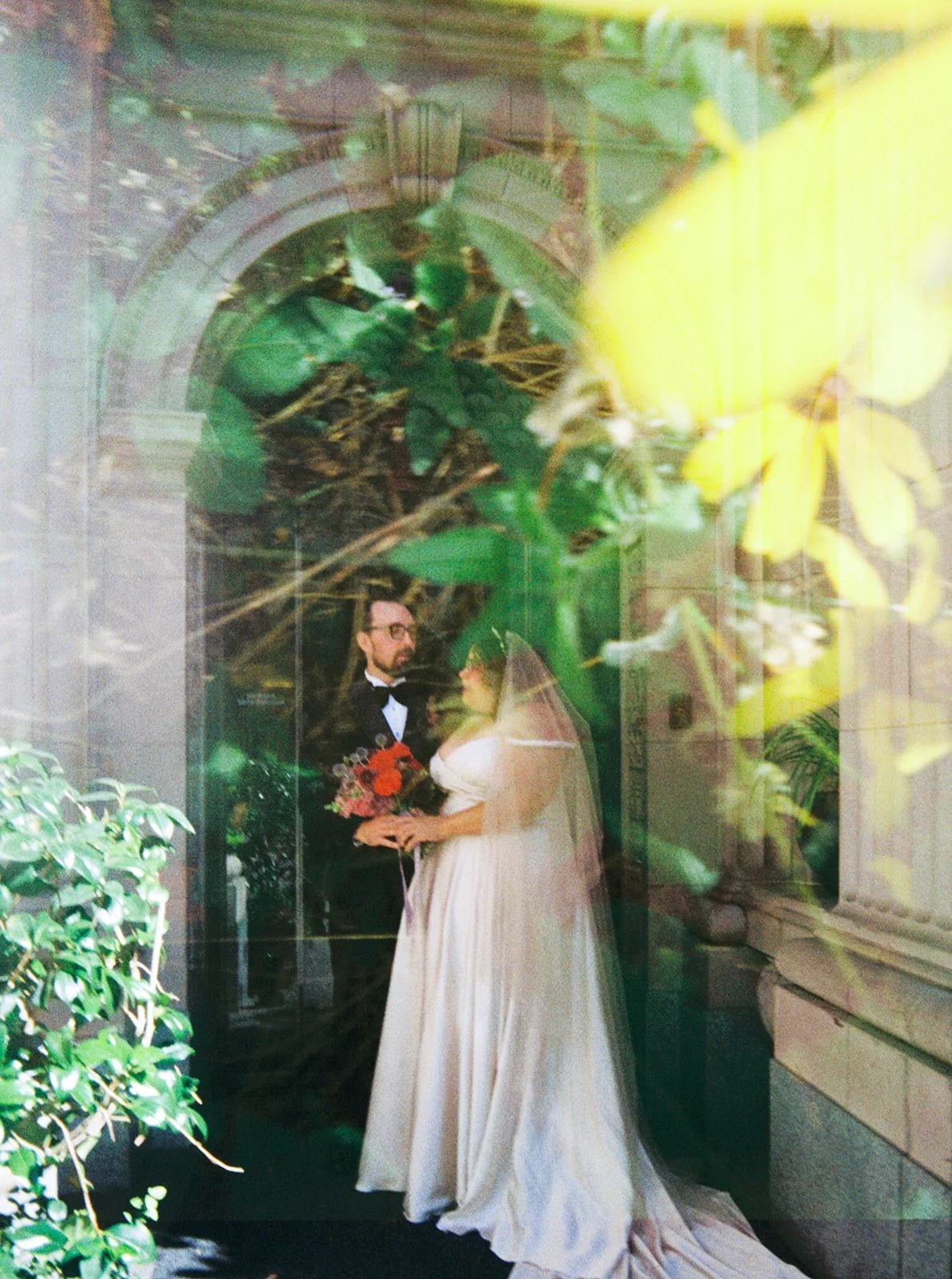 Bride and groom embracing outside Sentinel Hotel, partially framed by soft green foliage.