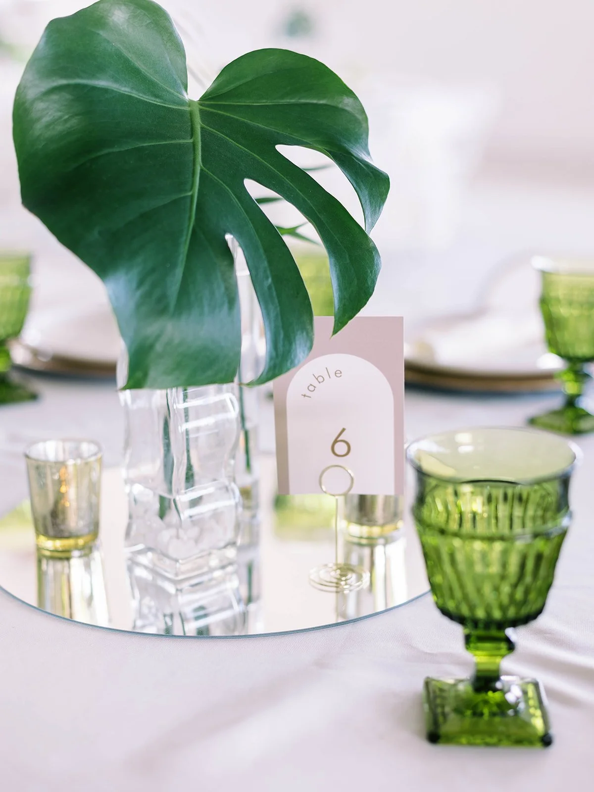 Close-up of a monstera leaf centerpiece in a crystal vase on a mirrored base, with a blush table number card and green goblet
