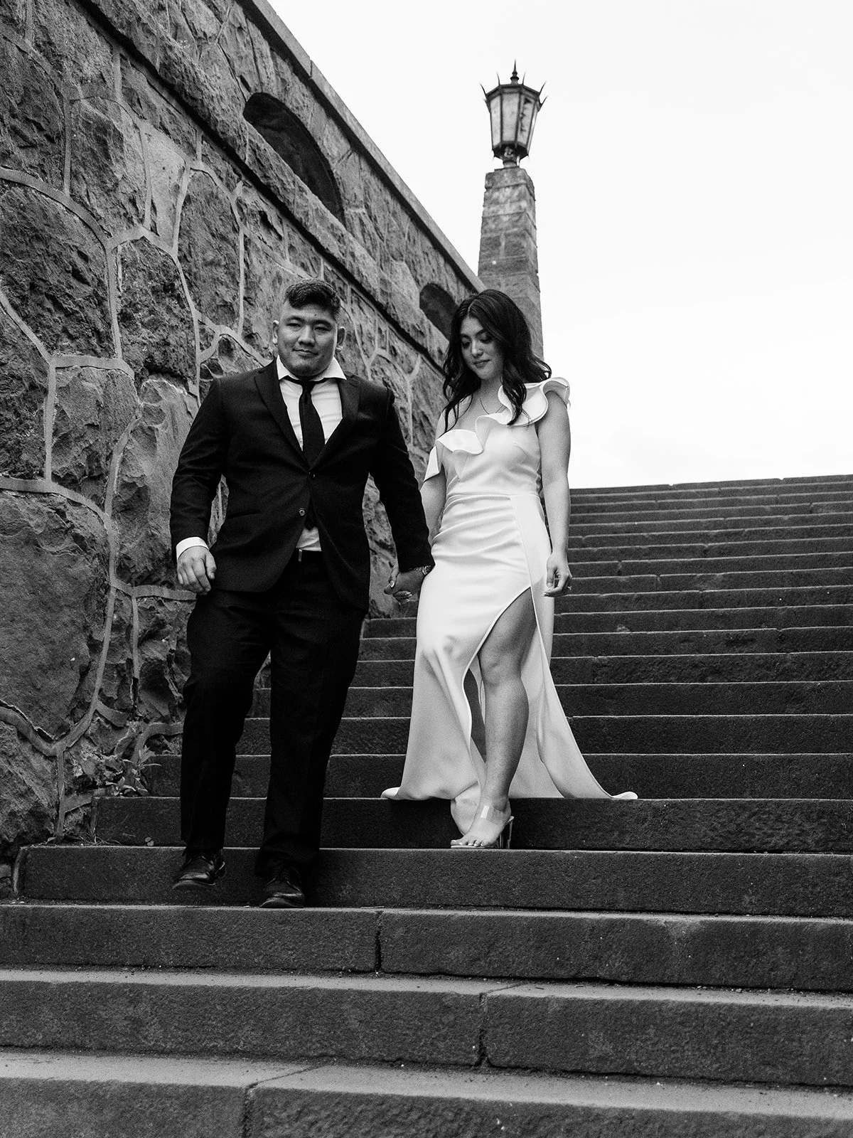 Black and white photo of the bride and groom walking hand-in-hand down stone stairs at The Grotto's upper overlook, a stone lamp post above them