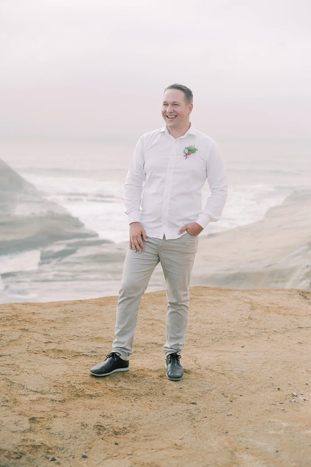 Groom standing on Oregon Coast cliff with boutonniere, ocean and rugged shoreline behind