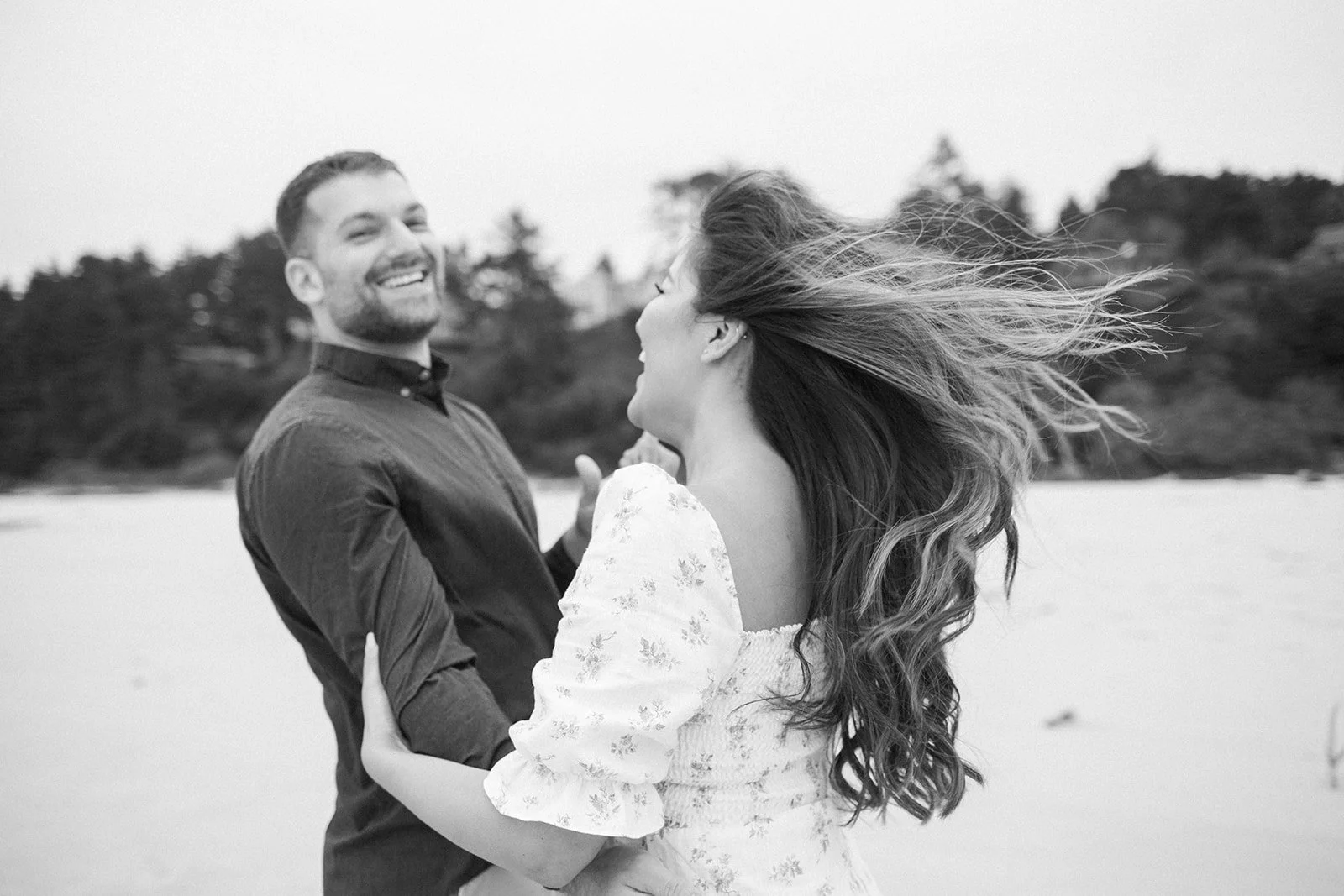 Couple laughing together on beach, woman’s hair blowing in wind, black and white coastal engagement photo
