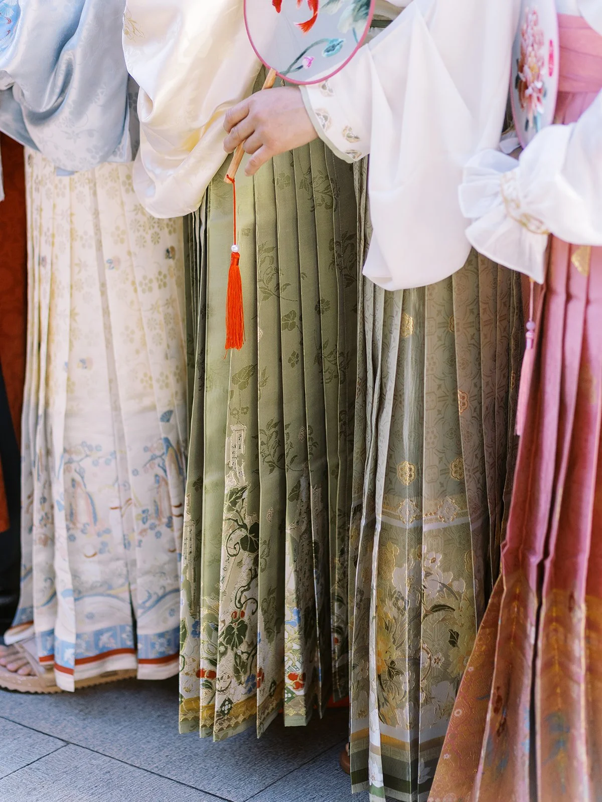 Close-up detail of layered hanfu skirts in sage green, blush pink, and ivory with embroidered patterns and a red tassel at a Chinese garden wedding