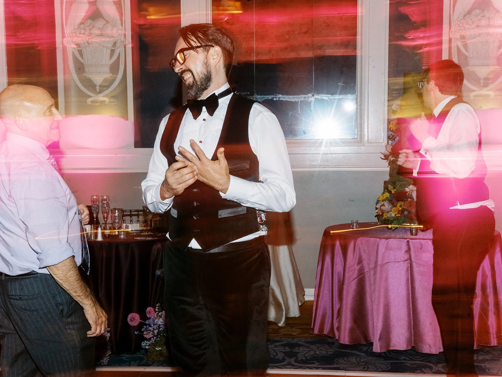 Groom laughing while talking with guest in reception space with pink light streaks and decorated tables.