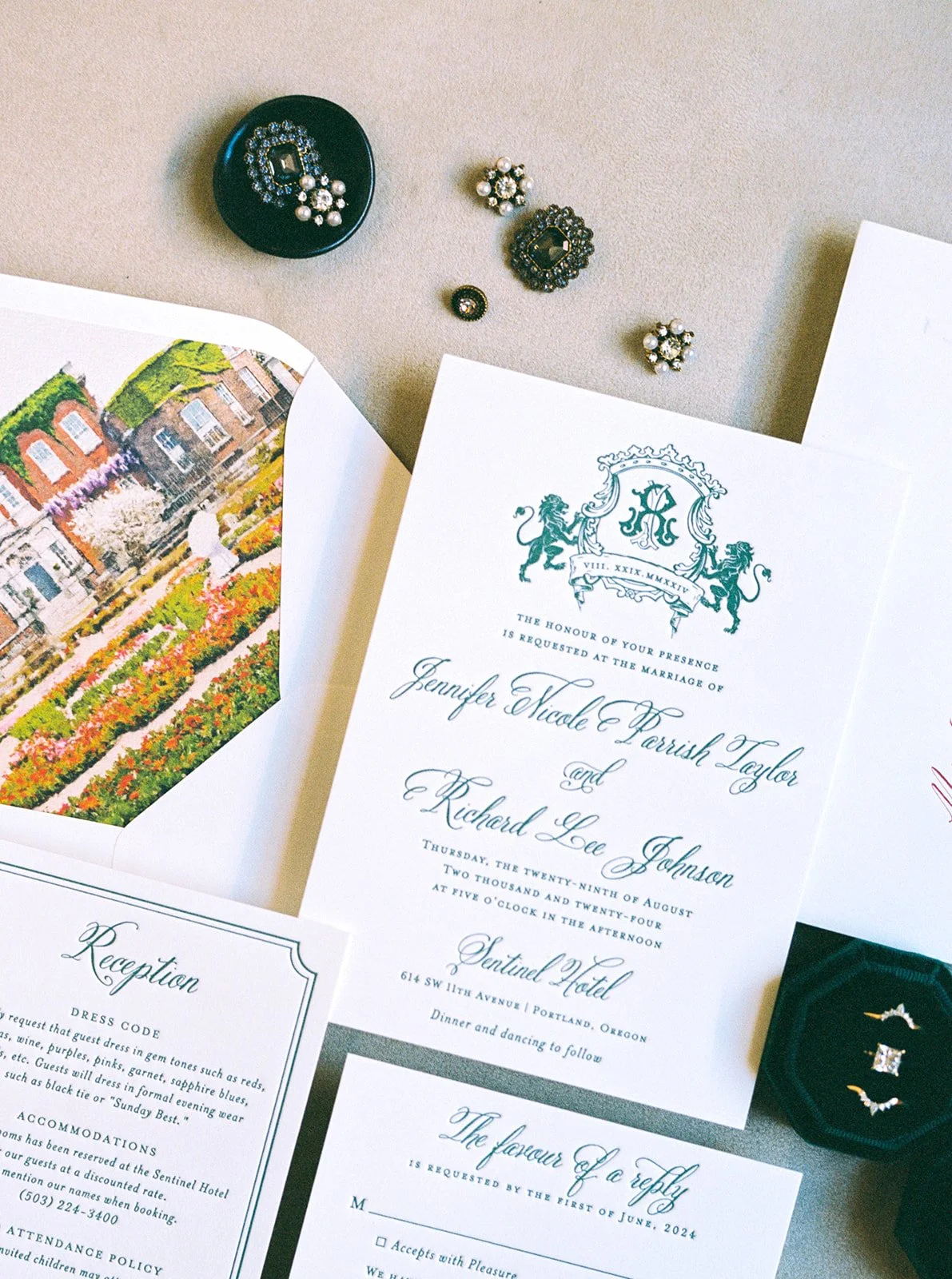 Elegant wedding invitation suite with crest design, styled with vintage brooches and rings on a neutral surface.
