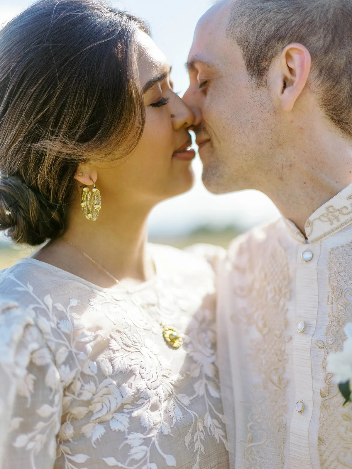 Close-up of bride and groom nearly kissing, bathed in warm sunlight, showcasing bride's gold earrings and embroidered gown details
