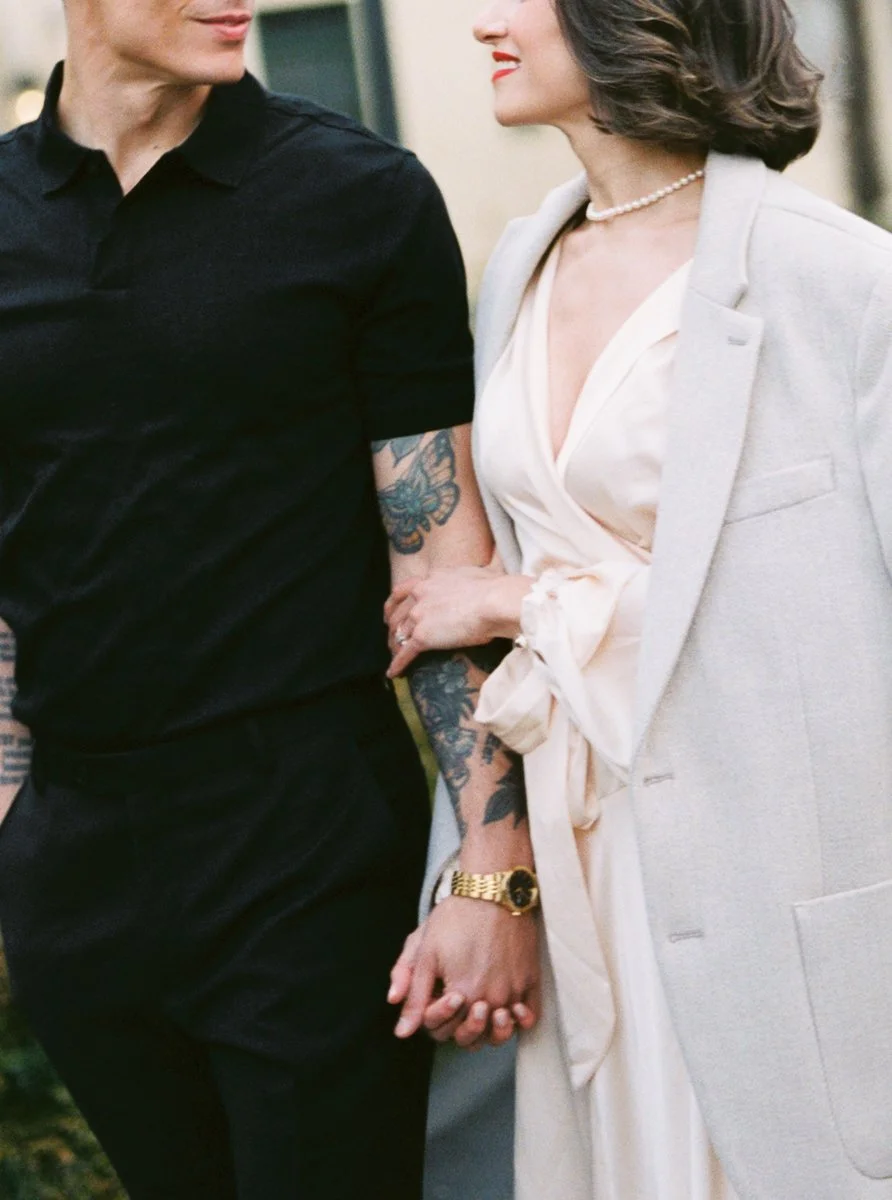 Close-up of couple holding hands, woman in cream dress and coat, man with tattoos in black outfit outdoors