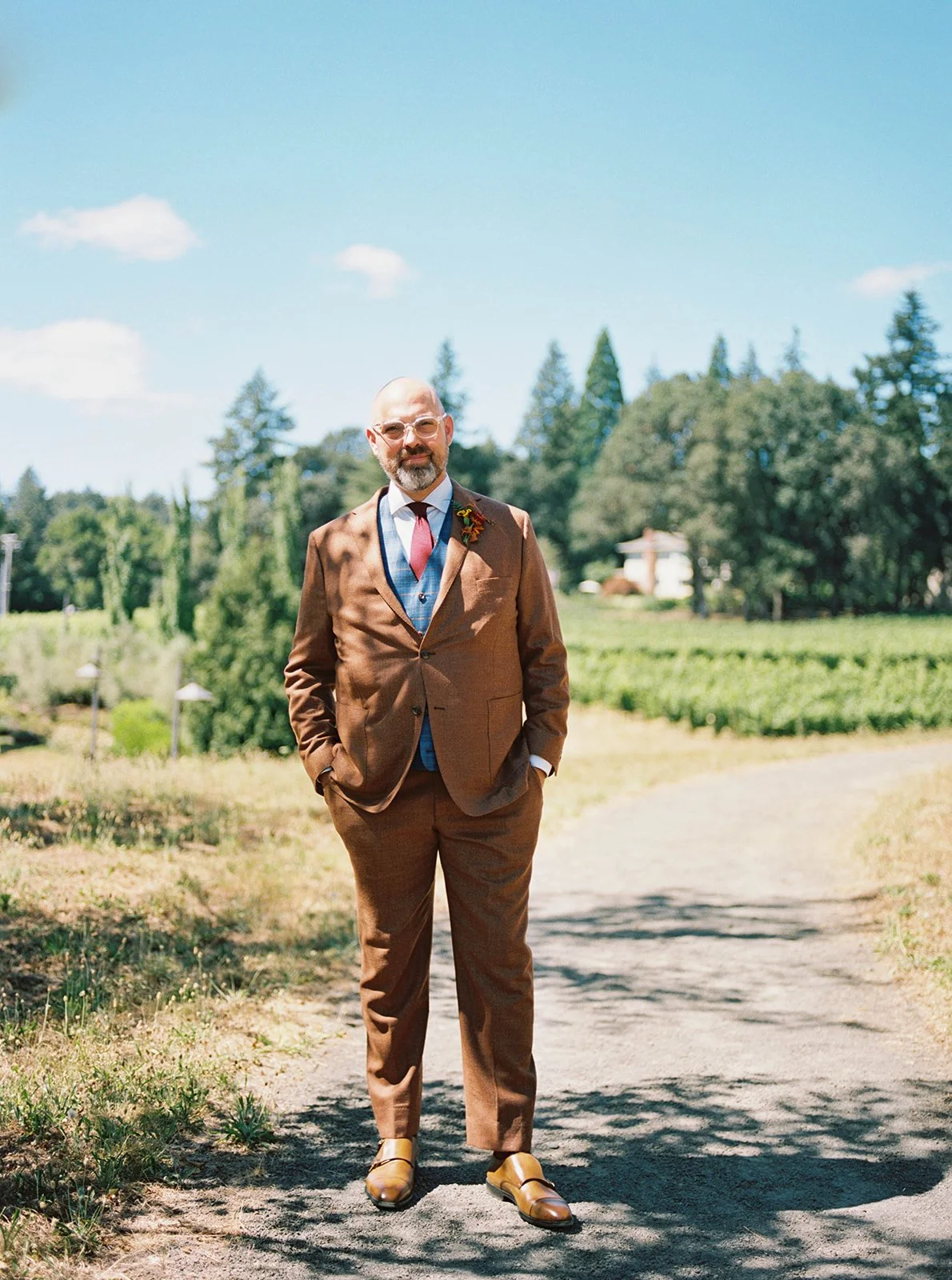Groom in brown suit standing on sunlit path with vineyard and trees in background