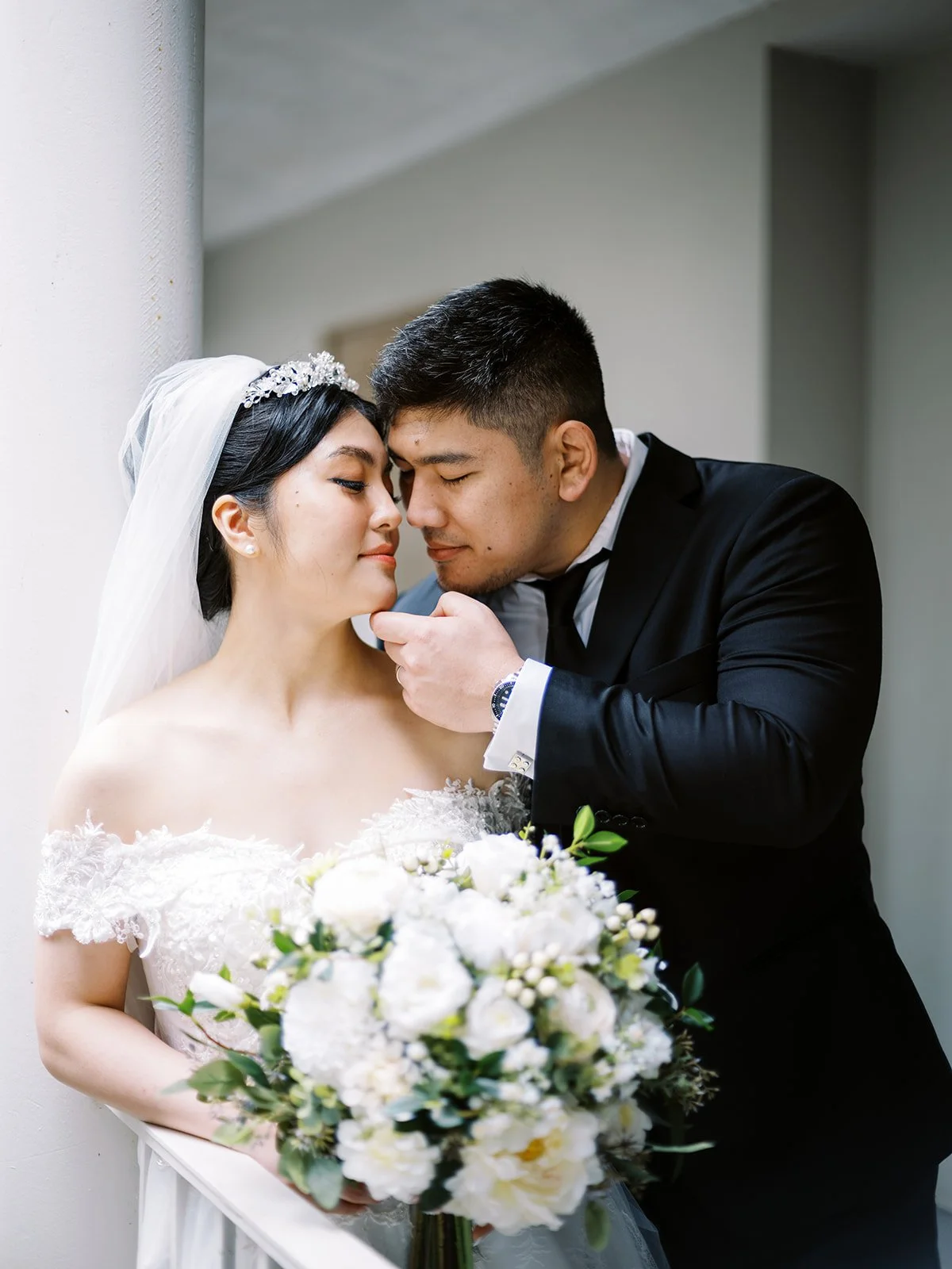 Close portrait of the groom with his hand on the bride's chin as she smiles with eyes closed on the colonnaded terrace at The Grotto, her white bouquet in the foreground