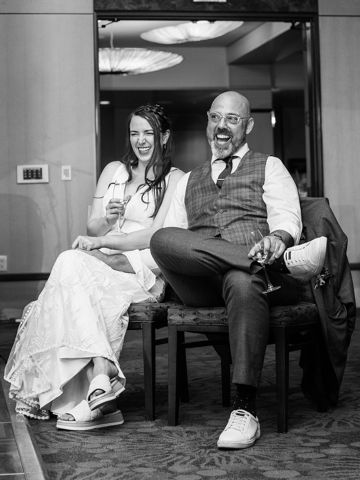 Black and white photo of the bride and groom laughing together in chairs during the reception, each holding a champagne glass