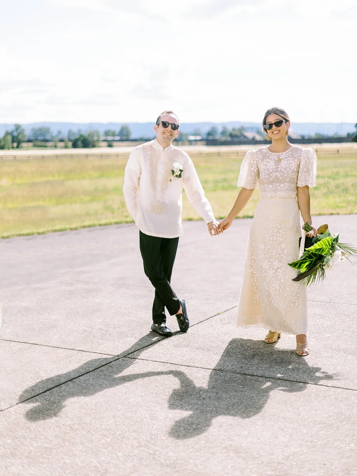 Bride and groom holding hands and smiling at the camera while walking on the Pearson Airfield tarmac, their shadows visible below
