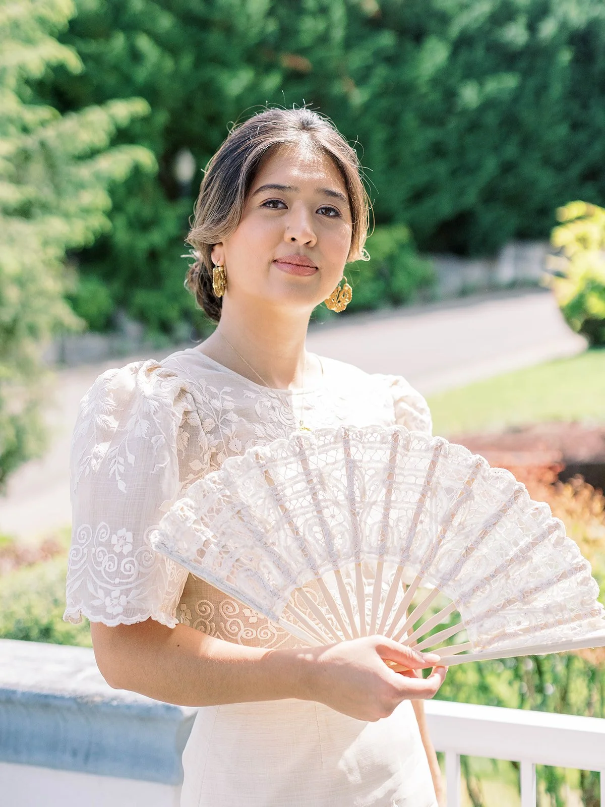 Bride in embroidered Filipino wedding dress holding a white lace hand fan, smiling softly in a garden setting

