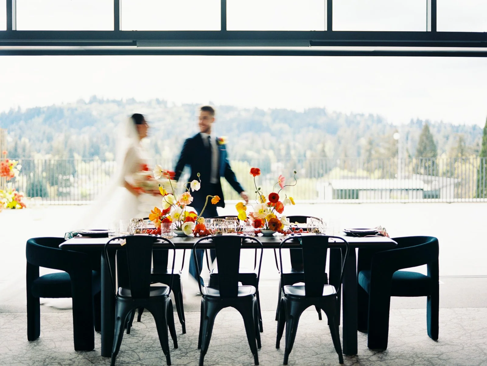 Bride and groom walk past a modern reception table with colorful floral centerpieces and large window views behind them.
