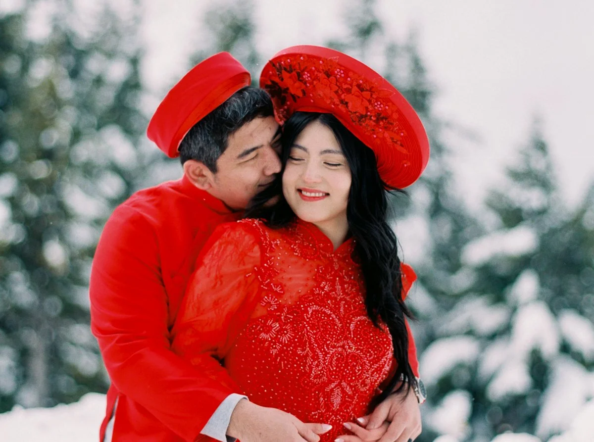 Couple embracing warmly in the snow, dressed in matching red áo dài and khăn đóng, surrounded by snowy pine trees.