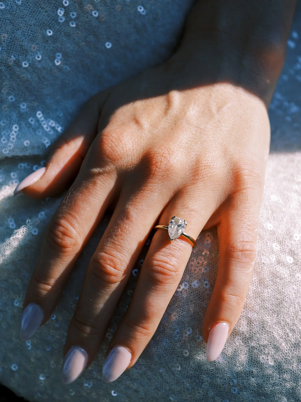  Close-up of a pear-shaped diamond engagement ring on a hand resting on a sequined skirt.