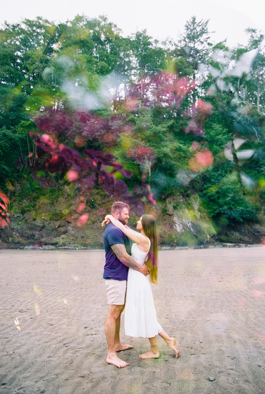 Couple standing barefoot on a beach, embracing with trees and cliffs in the background and soft floral overlay.