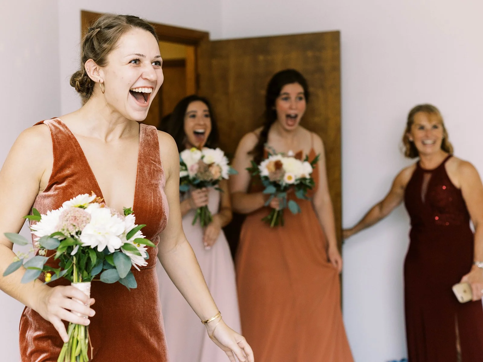 Bridesmaids react with surprise and smiles as they see the bride holding a bouquet in a getting-ready room.