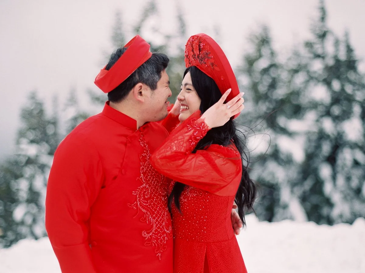 Couple in red Vietnamese áo dài smiling and adjusting headpieces in snowy Mount Hood forest.
