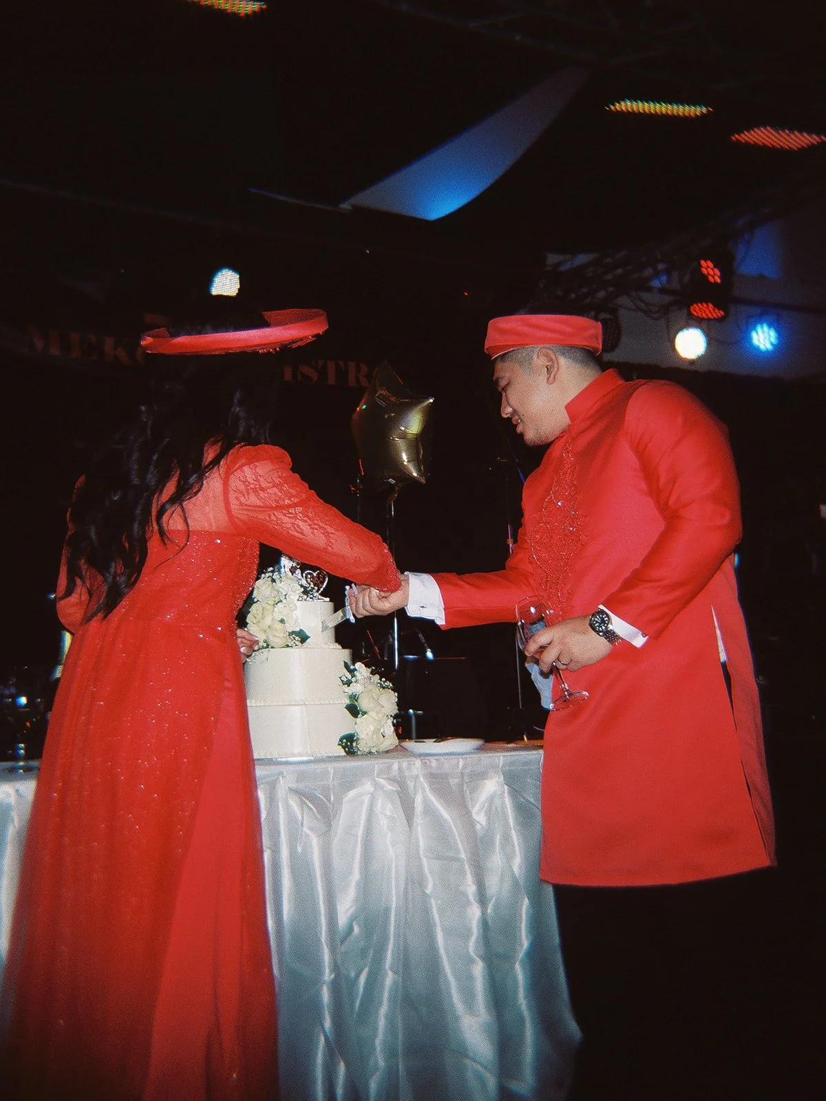 Bride and groom in red Vietnamese áo dài cutting their white tiered wedding cake together at Mekong Bistro, stage lights glowing above them