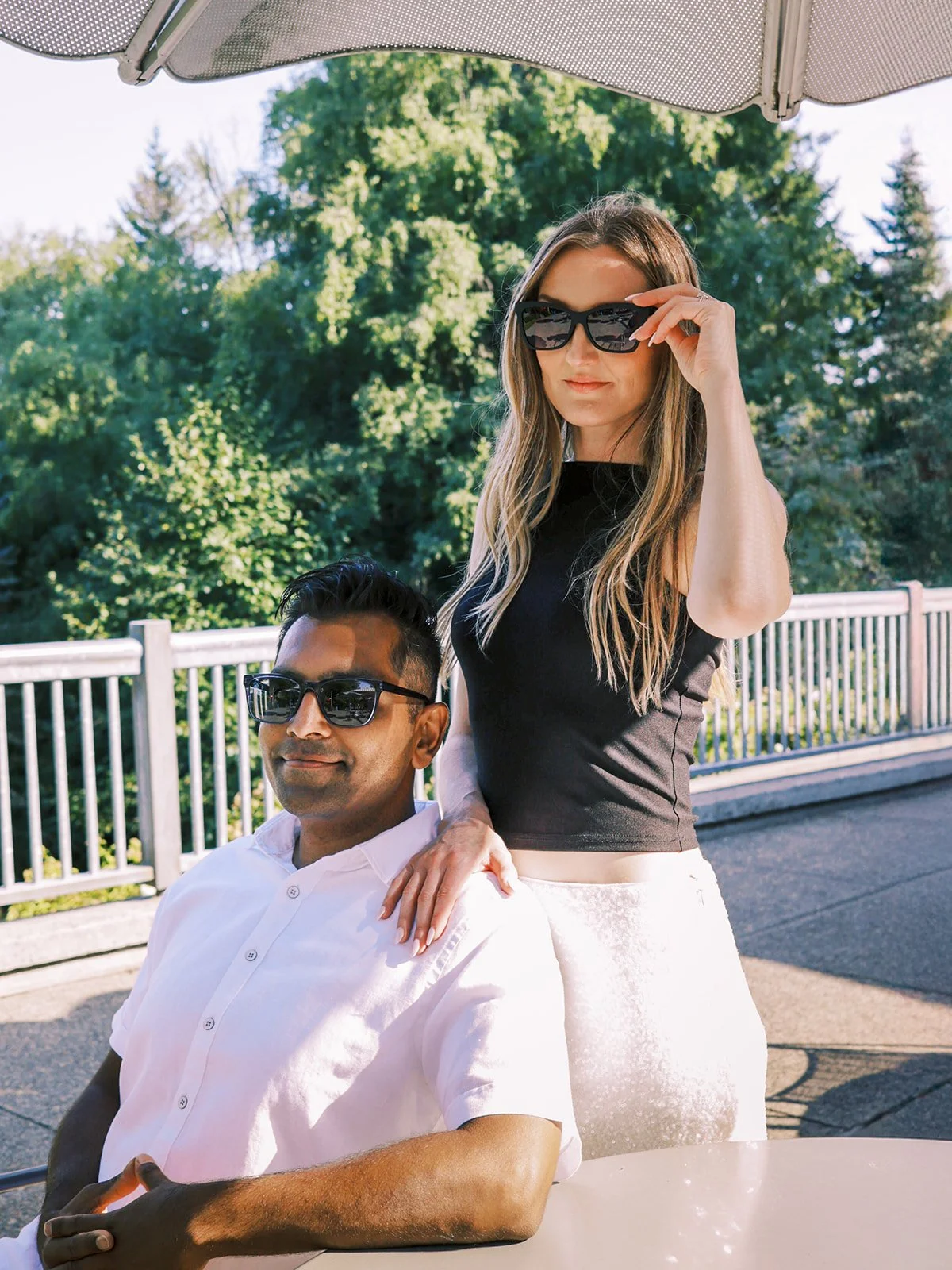 A couple in sunglasses posing under a patio umbrella outdoors — he seated at a table, she standing behind him.