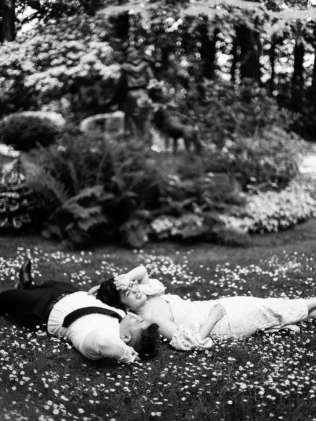 Black and white overhead shot of the bride and groom lying side-by-side on a daisy-covered lawn at The Grotto, laughing and looking up