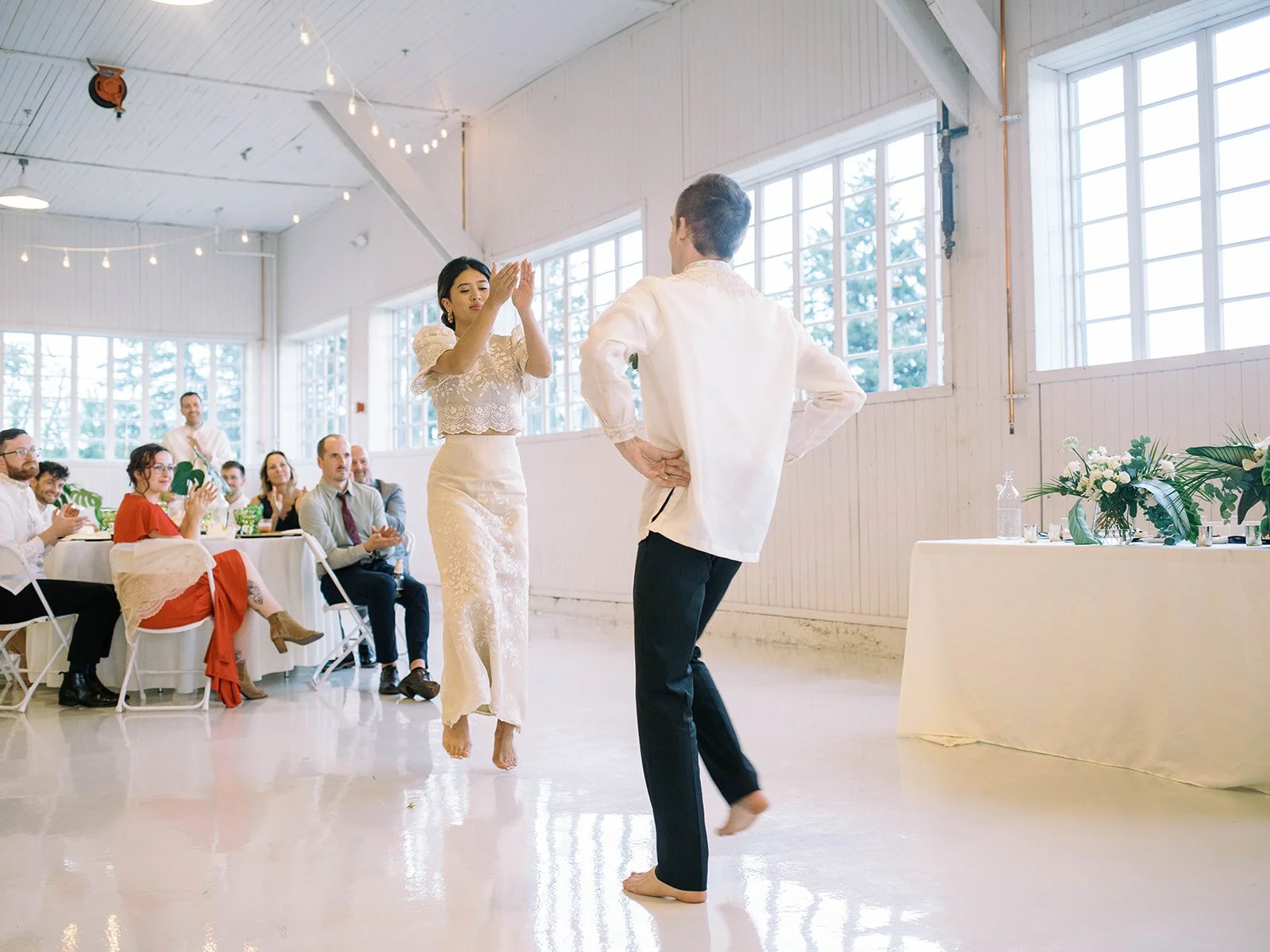 Bride and groom clapping hands barefoot during their choreographed first dance as delighted guests applaud at their reception tables
